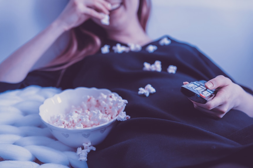 Smiling woman eating popcorn, with a bowl of popcorn beside her, and holding a television remote.