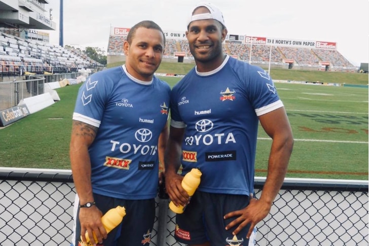 Two men in North Queensland Cowboys jerseys stand on beside the footy field holding yellow water bottles.