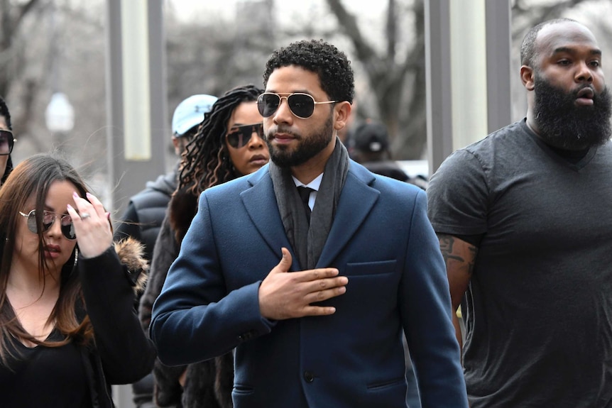 Jussie Smollett wears a blue suit and sunglasses and walks outside as he has several people behind and either side of him.