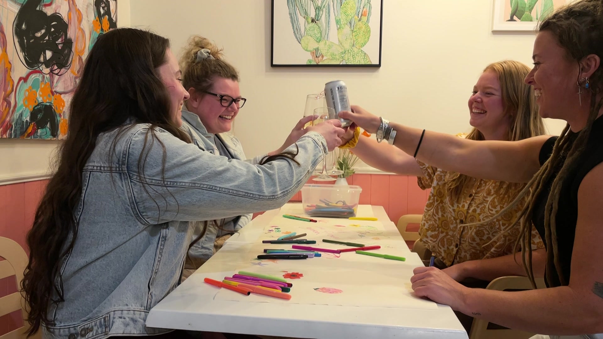 a group of four women make cheers at a table covered in colourful drawings and textas