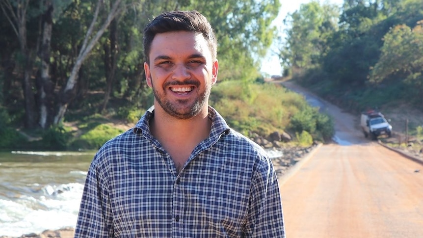 A man in a blue-check collared shirt standing near an outback river crossing, smiling.