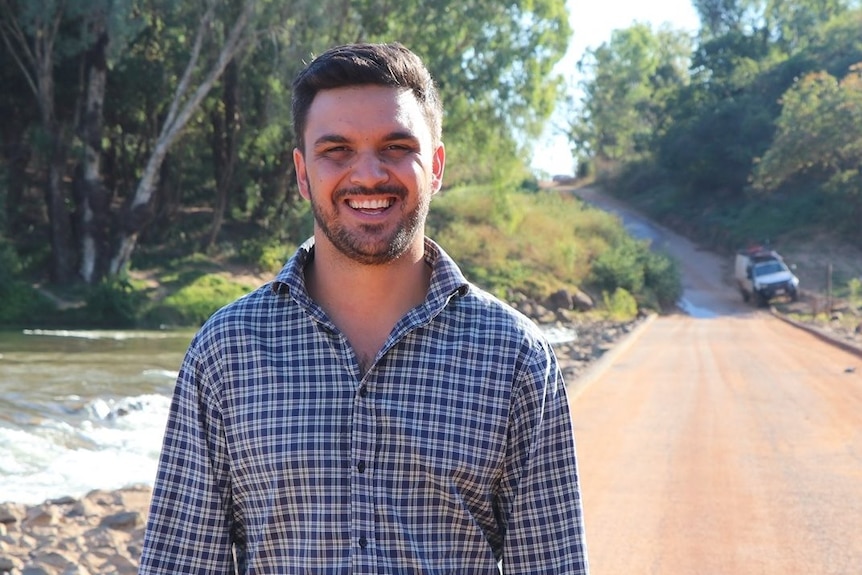 A man in a blue-check collared shirt standing near an outback river crossing, smiling.