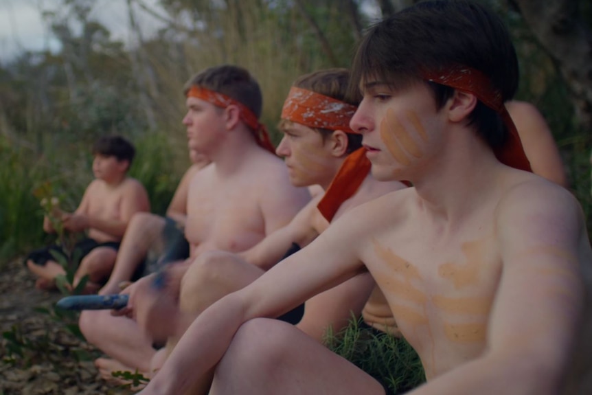 Four boys sit in a line in traditional Aboriginal dress.