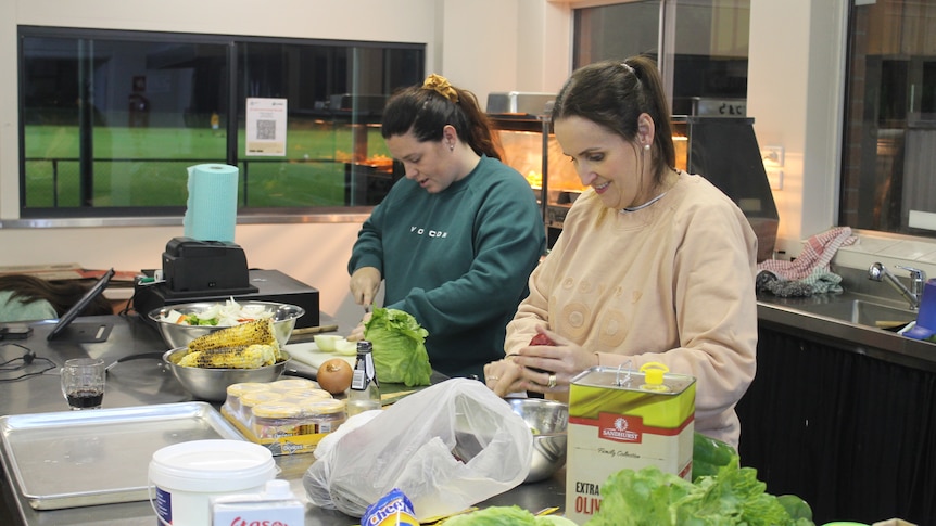 Two women prepare food in a commercial kitchen at a football club.