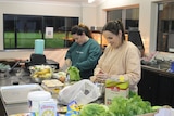 Two women prepare food in a commercial kitchen at a football club.
