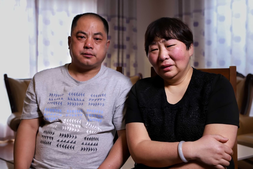 A man with short black hair and grey t-shirt, eyes bloodshot, sits next to a woman, eyes bloodshot, wearing black shirt.