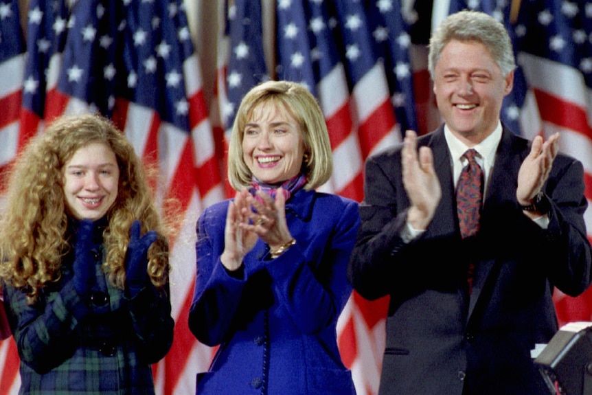 Chelsea, Hillary and Bill Clinton applauding in front of American flags.