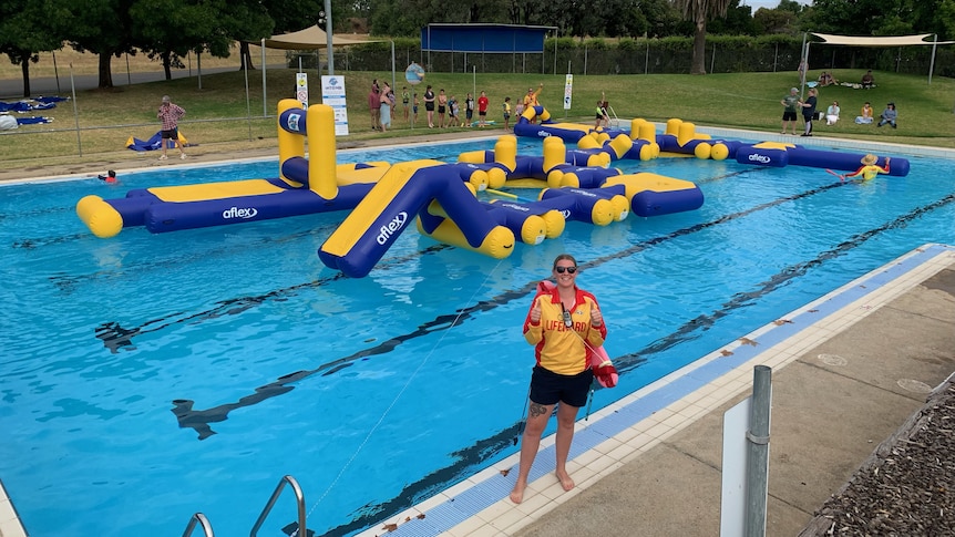a female lifeguard stands on the edge of an outdoor pool with a blue and yellow inflatable obstacle course on the water behind