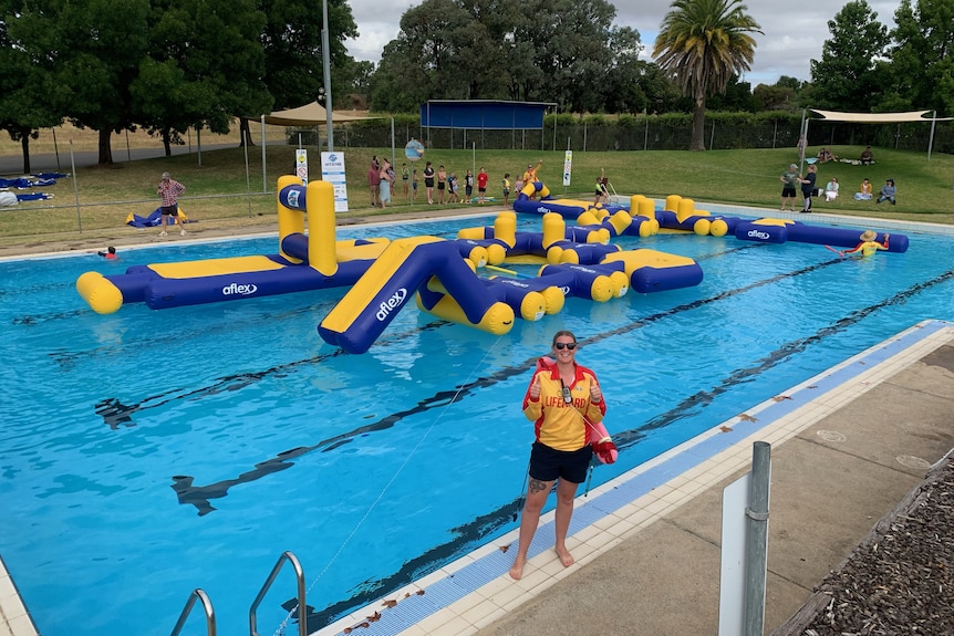 a female lifeguard stands on the edge of an outdoor pool with a blue and yellow inflatable obstacle course on the water behind