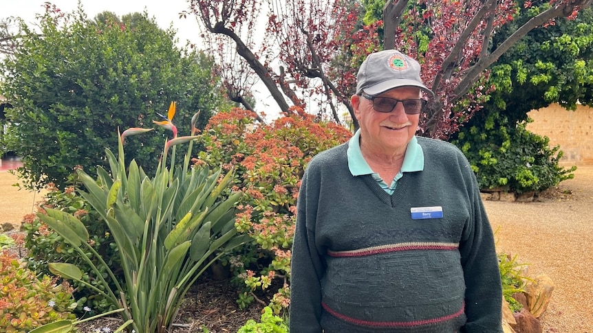 A man in a grey jumper and cap, wearing sunglasses and standing in front of a garden bed