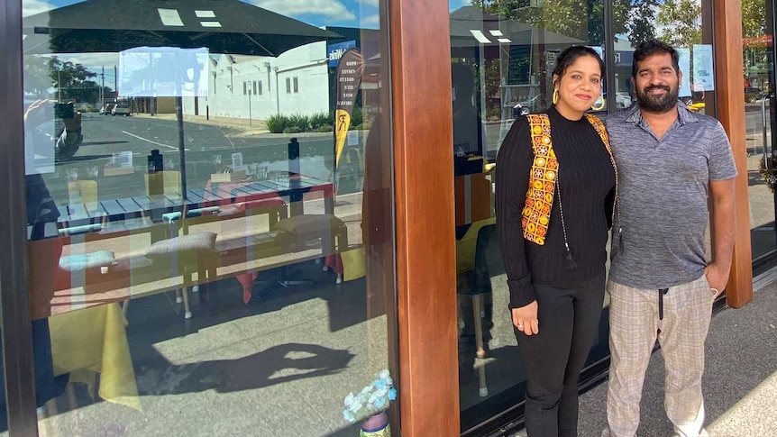 A man and a woman stand in front of a restaurant with glass walls. There is a sign that says 'Rooh' on the glass.