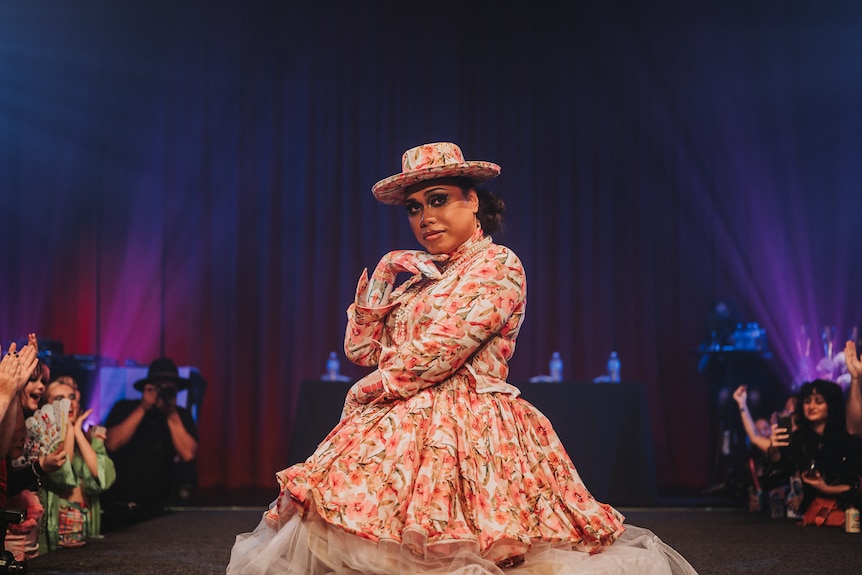 Neesha Alexander stands at centre stage in an peach coloured floral dress and hat. 