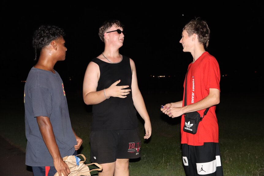 Three young men talking to one another and smiling while standing on a beach at night.