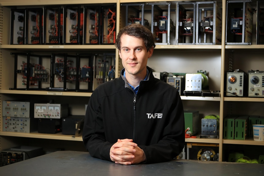 Matt Gallina stands with his hands folded in front, resting on a table. He stands in front of electrical items in a classroom.