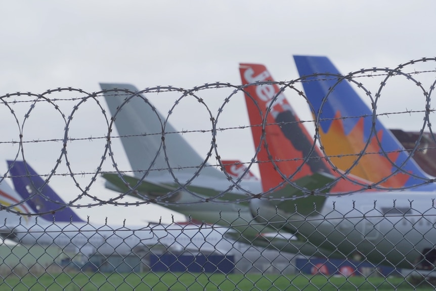A group of planes sit behind a barbed wire fence on the tarmac.