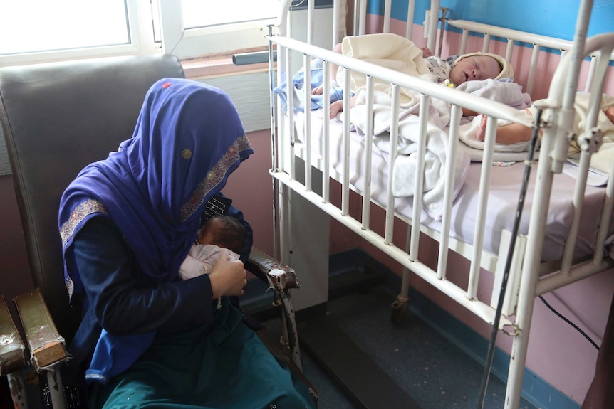 A mother breastfeeds her two-day-old baby as another infant lies in a crib nearby.