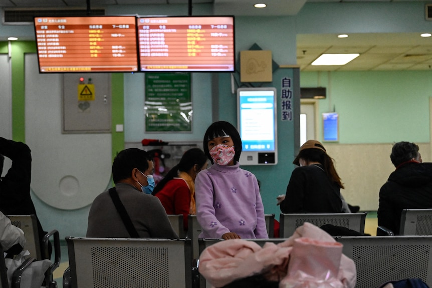 A Chinese girl wearing a mask stands beside an adult in a waiting room.