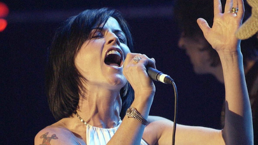 Dolores O'Riordan holds a microphone while she sings on stage.