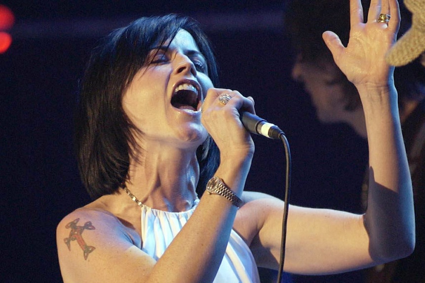 Dolores O'Riordan holds a microphone while she sings on stage.