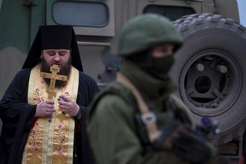 A priest wearing black robes, a gold stole and holding a cross, prays near a soldier and tank