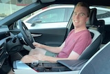 Young man sits in new car at the dealership smiling at camera.