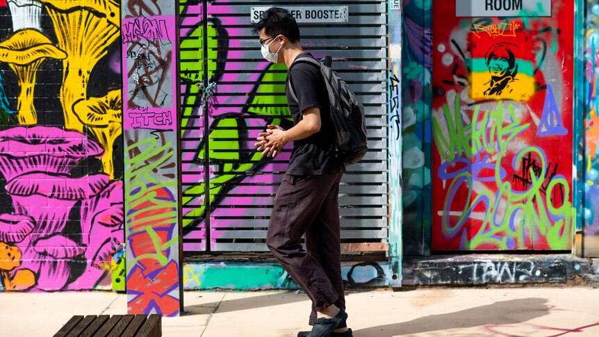 A man wearing a mask walking past a wall of colourful graffiti