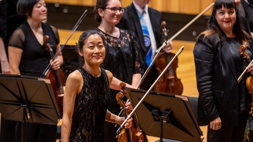 Violinist Natsuko Yoshimoto standing in front of the orchestra smiling at the audience 