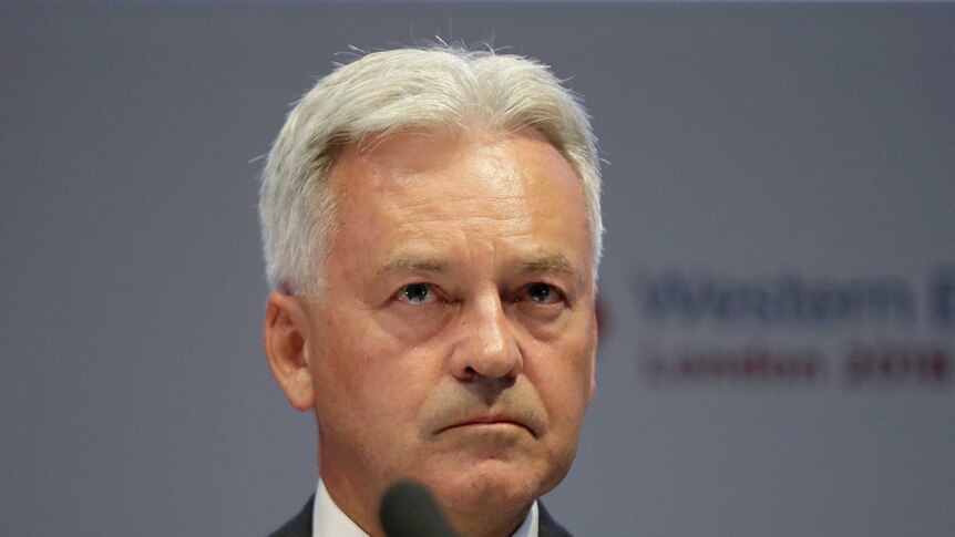 Alan Duncan sits behind a microphone as he stares blankly ahead in dark suit and light blue tie.