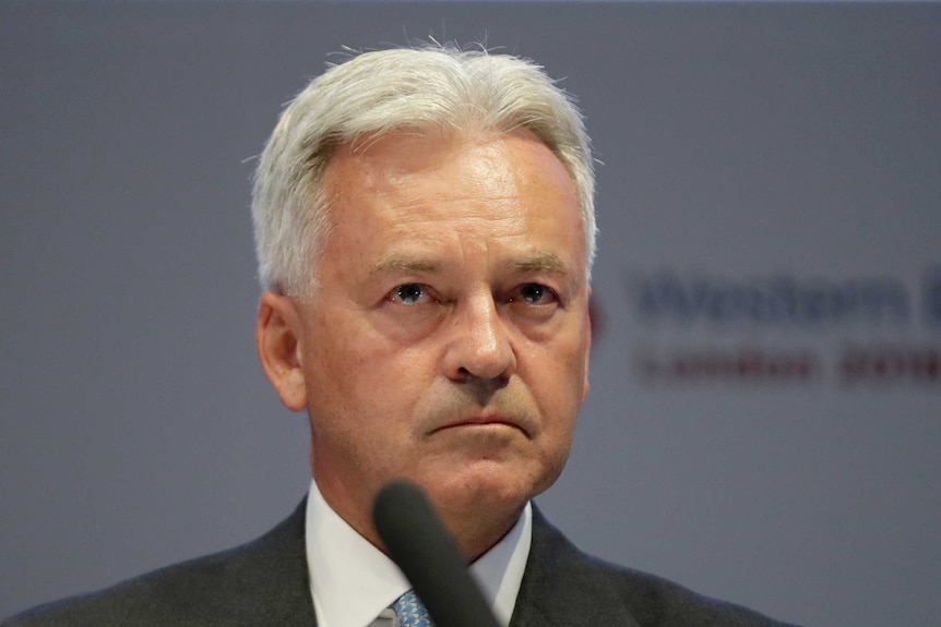 Alan Duncan sits behind a microphone as he stares blankly ahead in dark suit and light blue tie.