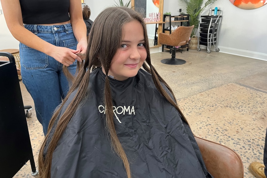 A young girl smiling with a covering over her clothes having her long hair plaited.