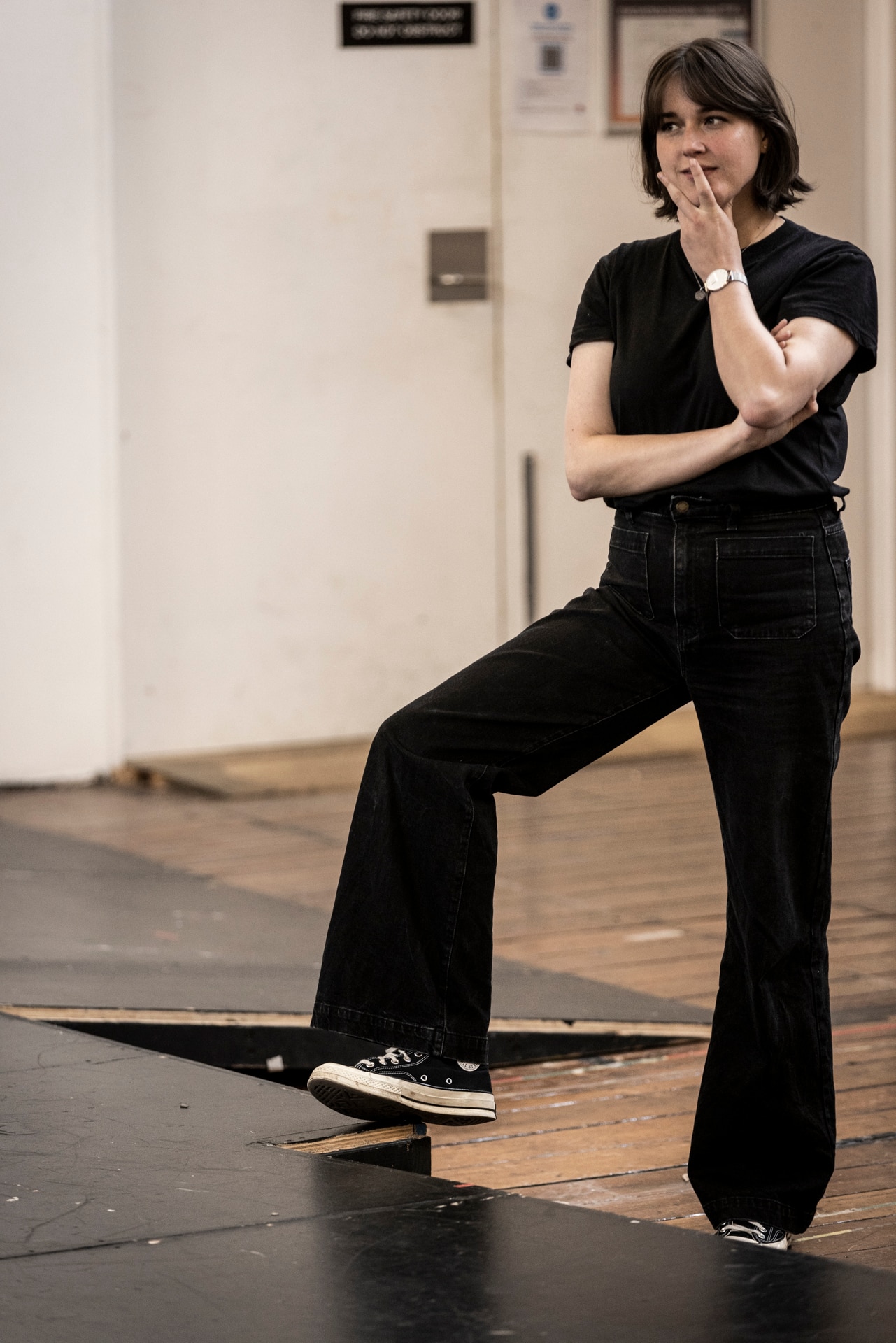 A woman with brown cropped hair and bangs stands with her hand on her chin, her foot resting on a makeshift stage