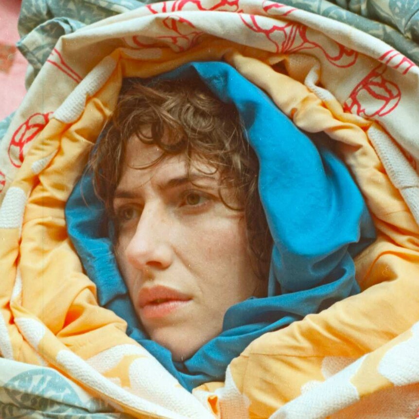 Aldous Harding is a New Zealand singer. She is wrapped up in multiple blankets and looking sideways.