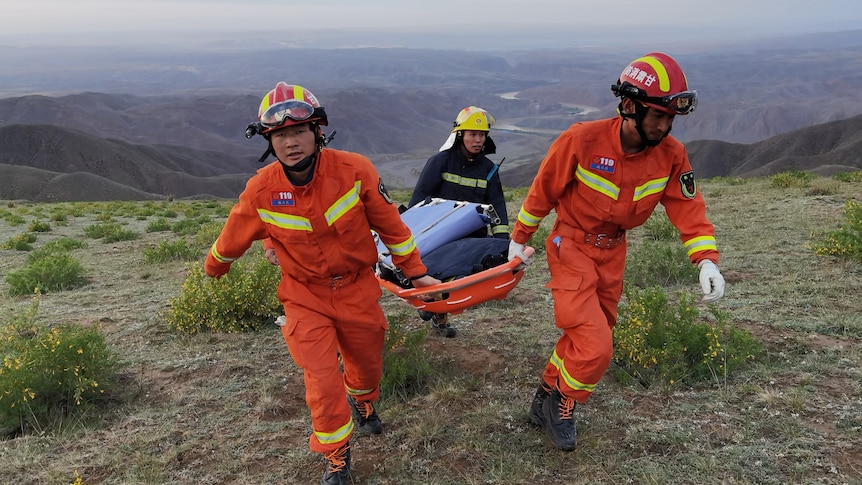 Men in red search and rescue uniforms carry a stretcher on top of a mountain. 