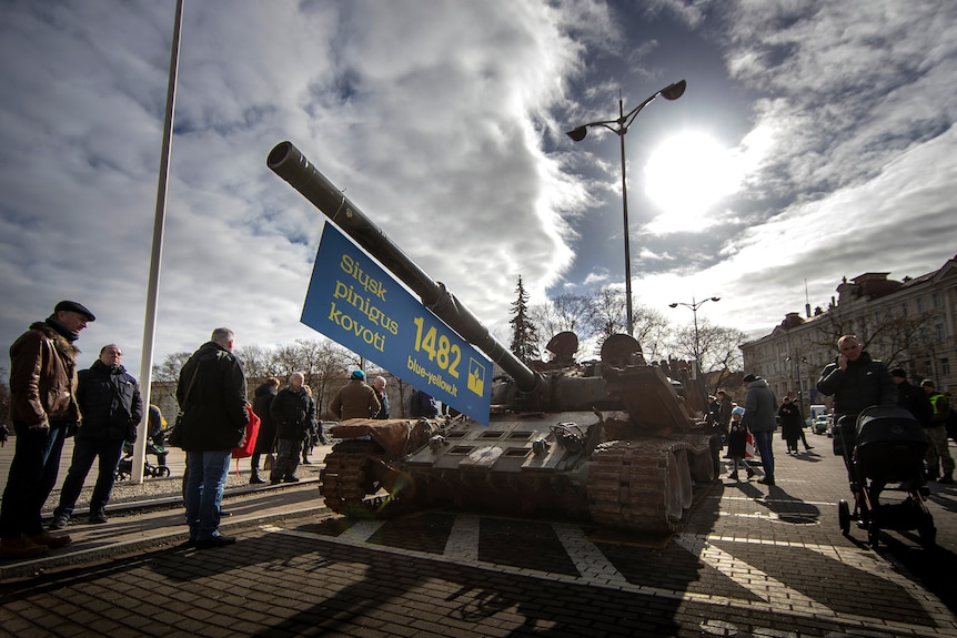 People walk past Russian tank on display, decorated with blue and yellow banner reading "Send money to fight".