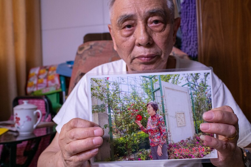 An old Chinese man holds up a picture of a middle-aged woman in a garden.