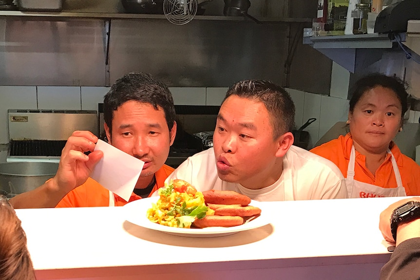 Two men look at a restaurant docket across a kitchen bench.