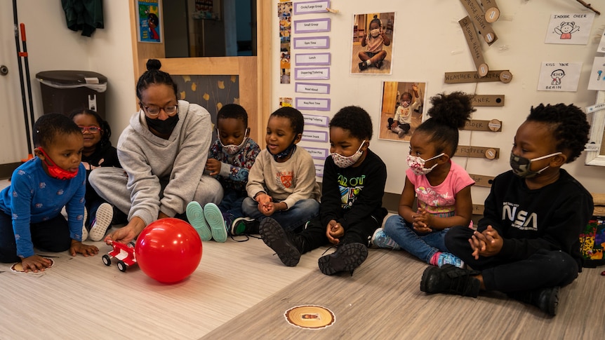 Children sit on the floor while a childcare worker pushes a toy truck