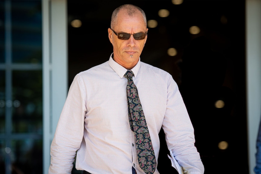 A man wearing a white shirt walks outside a court room. He has a black tie.