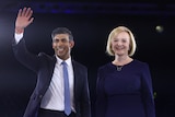 Rishi Sunak, wearing a suit and blue tie, waves as he stands next to Liz Truss, who wears a navy dress.