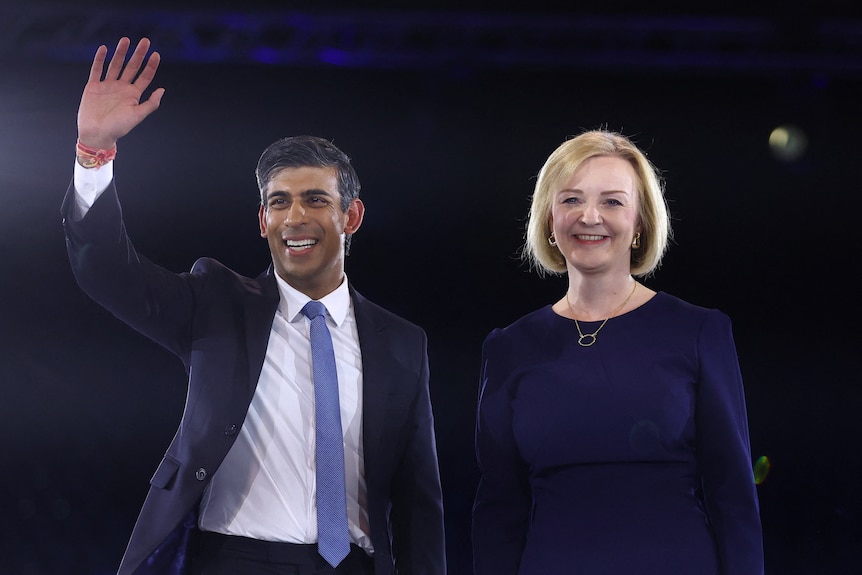 Rishi Sunak 身穿蓝色西装，打着领带，站在身穿海军蓝连衣裙的 Liz Truss 旁边挥手致意。