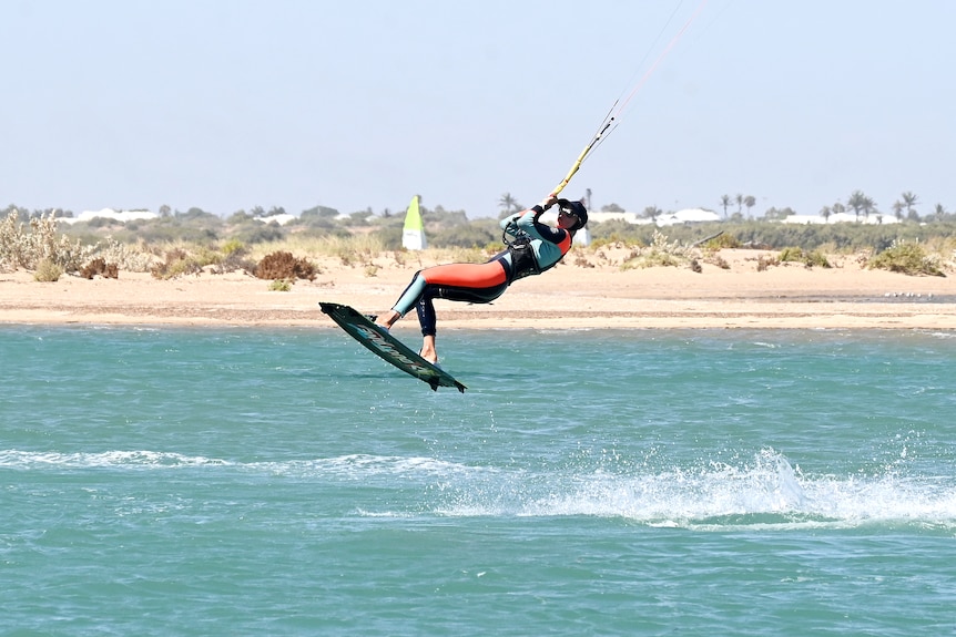 A  kiteboarder in the middle ground starts an aerial trick over blue waters with an arid landscape in the background.