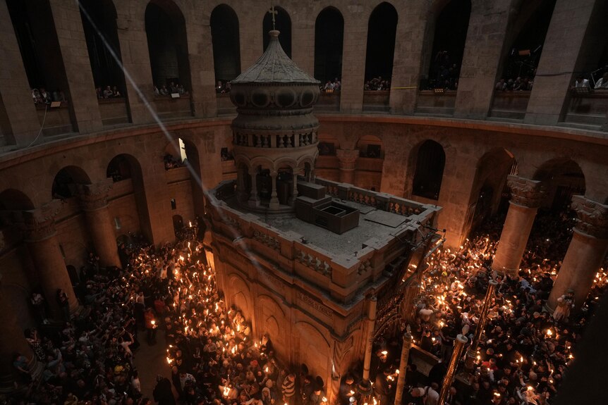 A view from above of people gathering inside holding candles which give off a bright orange glow around the old architecture.