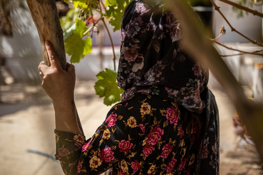 A photo of Rahima, who wears a hijab, shown from the back, holding on to a tree.