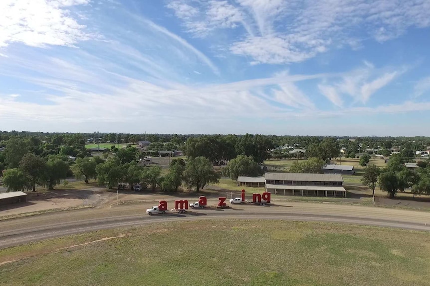 Aerial shot of red sculptural letters on a back of three utes being towed around a country town