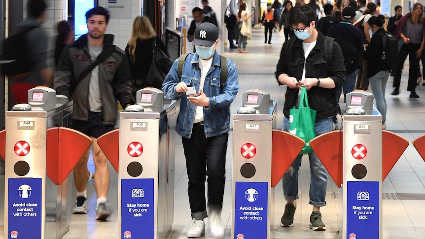a woman and a man wearing mask covering their nose and mouth walking through the opal card readr at a train station