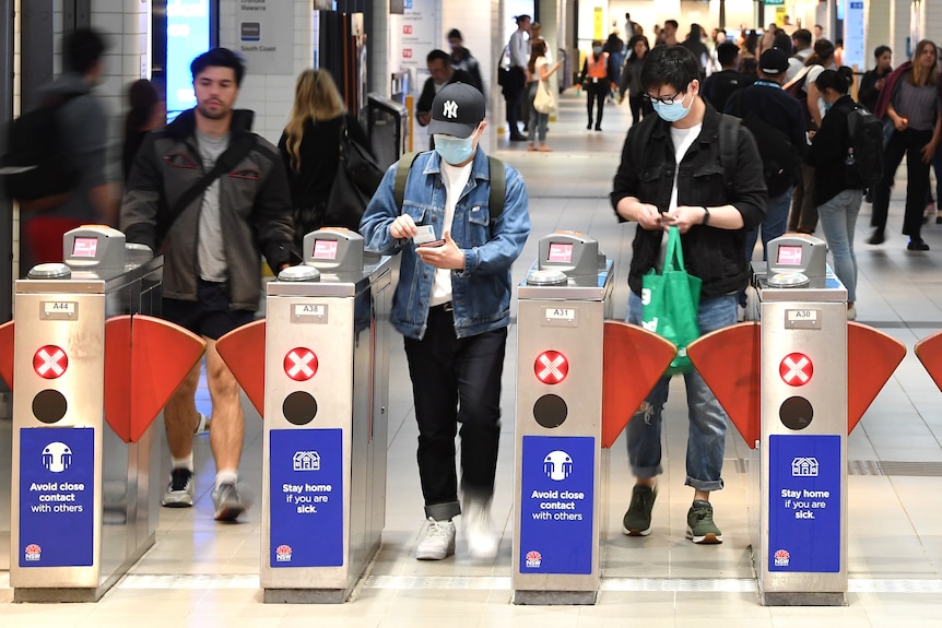 a woman and a man wearing mask covering their nose and mouth walking through the opal card readr at a train station