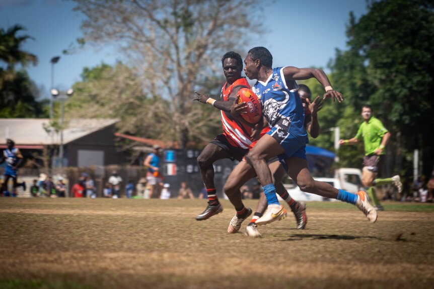 Two men, one wearing red and one wearing blue, run on a field. The player in blue holds a football.