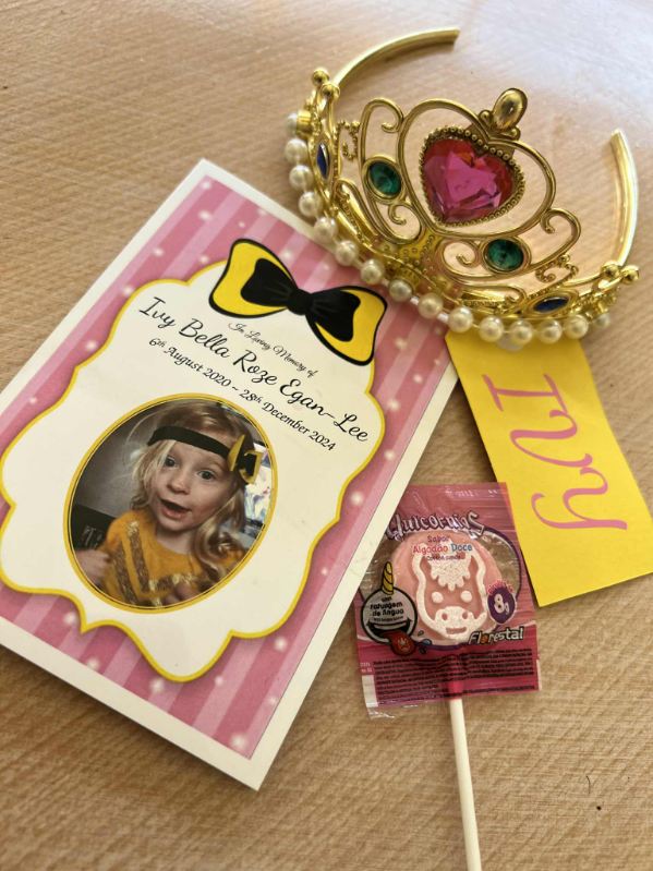 Un folleto funerario rosa y amarillo con la imagen circular de una niña rubia y una etiqueta amarilla con su nombre que dice 