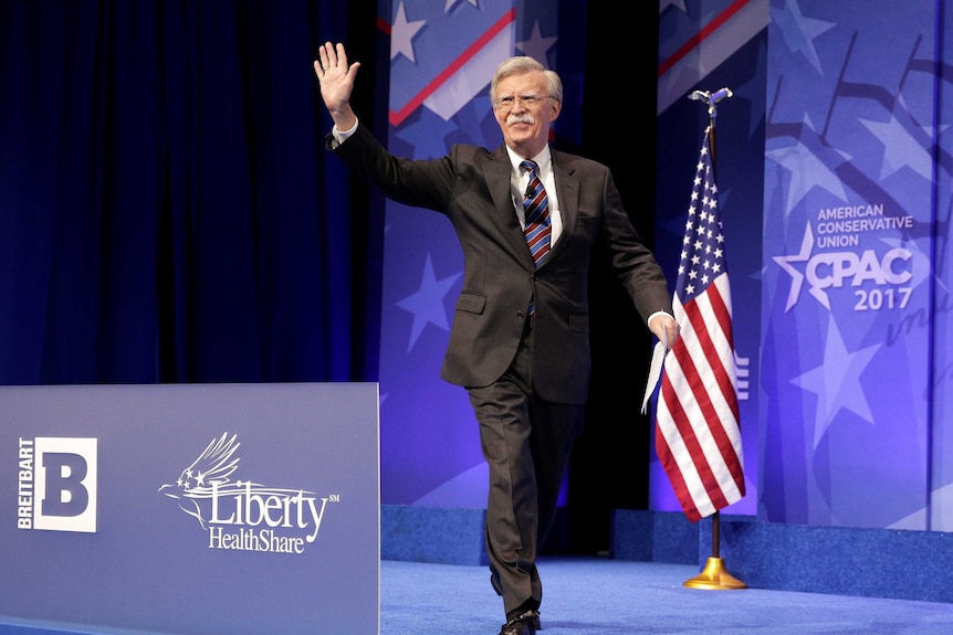 A man in a dark suit waves to a crowd while walking towards a lectern.