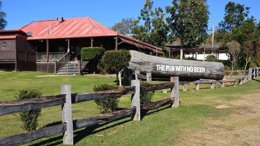 A large building, with red tin sloping roof,  stairs, wrap-around verandah, large sign on log, "Pub with no beer", wooden fence.
